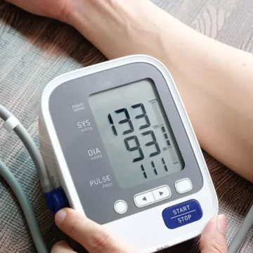 Uma pessoa medindo a pressão arterial em casa com um aparelho digital, sentada corretamente com os pés no chão e o braço na altura do coração.