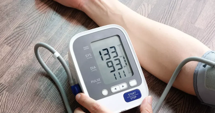 Uma pessoa medindo a pressão arterial em casa com um aparelho digital, sentada corretamente com os pés no chão e o braço na altura do coração.
