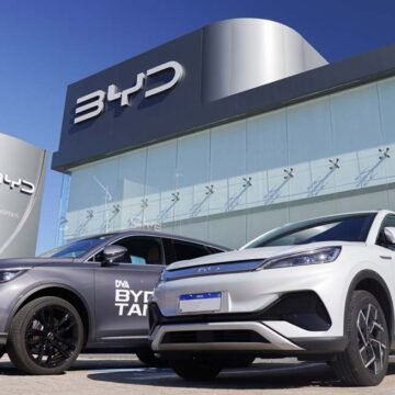 Uma imagem de um carro BYD em destaque em uma concessionária brasileira, com placas de preço e banners de promoções ao fundo.