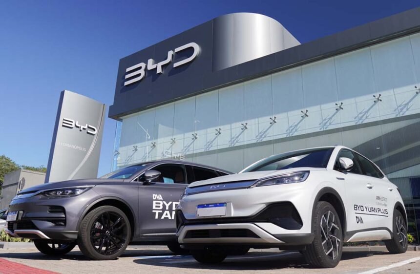 Uma imagem de um carro BYD em destaque em uma concessionária brasileira, com placas de preço e banners de promoções ao fundo.