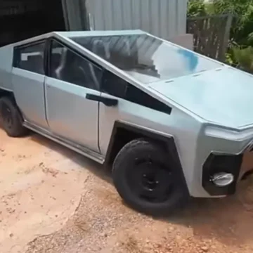 Lucas Carvalho posa ao lado de seu CyberFusca prateado, um carro com design futurista construído sobre um chassi de Fusca. O carro tem linhas retas e angulares, e uma caçamba na parte traseira.