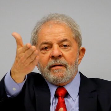 Foto de Lula em um evento, cumprimentando empresários em um ambiente formal, com um fundo de bandeiras do Brasil
