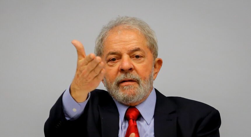 Foto de Lula em um evento, cumprimentando empresários em um ambiente formal, com um fundo de bandeiras do Brasil