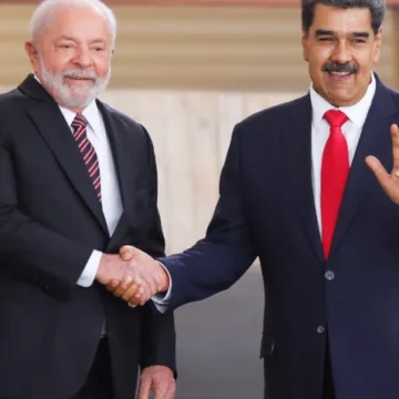 Uma sala de reunião oficial, com Nicolás Maduro ao centro, gesticulando enquanto fala para um público atento.