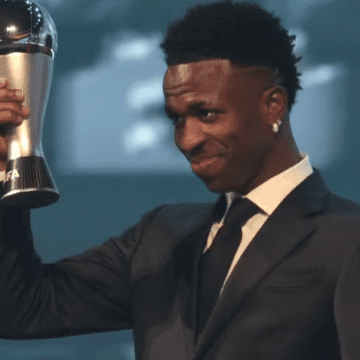 Vinícius Júnior comemorando sua vitória como Melhor Jogador do Mundo em 2024, com a taça da Fifa em mãos, no palco da cerimônia.