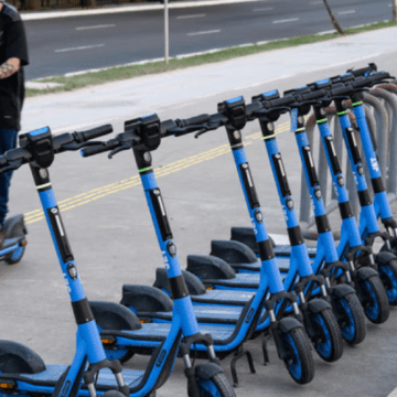 Patinete elétrica estacionada em uma estação compartilhada no bairro de Pinheiros, com pedestres e ciclistas ao fundo.