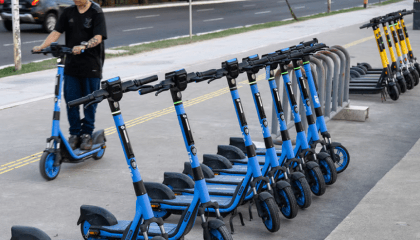 Patinete elétrica estacionada em uma estação compartilhada no bairro de Pinheiros, com pedestres e ciclistas ao fundo.