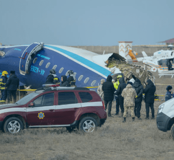 Destroços de um avião com a cauda danificada em um campo, com partes da fuselagem espalhadas.