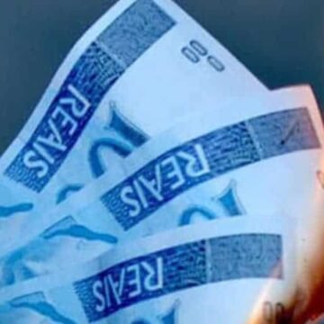 Cédulas de dólar espalhadas sobre uma mesa de madeira, ao lado de gráficos financeiros digitais mostrando flutuações da moeda americana.