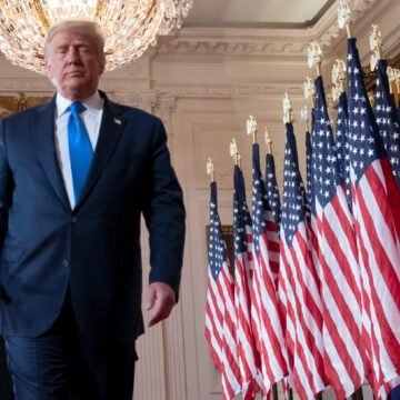Donald Trump em um palco, acenando para apoiadores, com bandeiras dos EUA ao fundo.