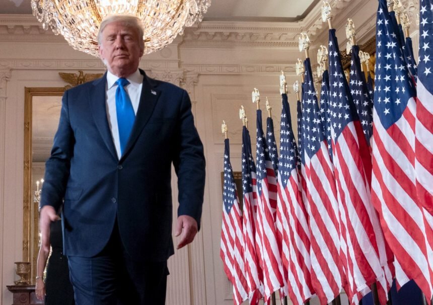 Donald Trump em um palco, acenando para apoiadores, com bandeiras dos EUA ao fundo.