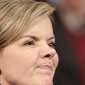 Gleisi Hoffmann, presidente do PT, em um momento de reflexão, com o logotipo do partido ao fundo, simbolizando a possibilidade de sua candidatura à Presidência em 2026.