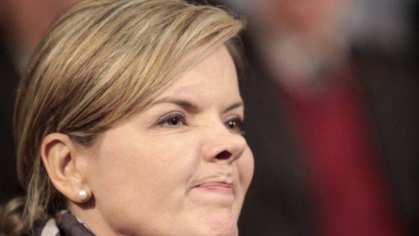 Gleisi Hoffmann, presidente do PT, em um momento de reflexão, com o logotipo do partido ao fundo, simbolizando a possibilidade de sua candidatura à Presidência em 2026.