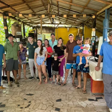 Uma imagem horizontal que mostra os membros da família Bettim em meio à plantação de café, em sua fazenda. Eles estão em grupos, com alguns olhando para a câmera e outros trabalhando. A cena transmite a conexão da família com a terra e a dedicação ao trabalho.